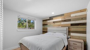 A bedroom with a bed, a nightstand, a window, and a wooden wall panel. - Coastal 2-Bedroom Home Minutes to Beach (Englewood)