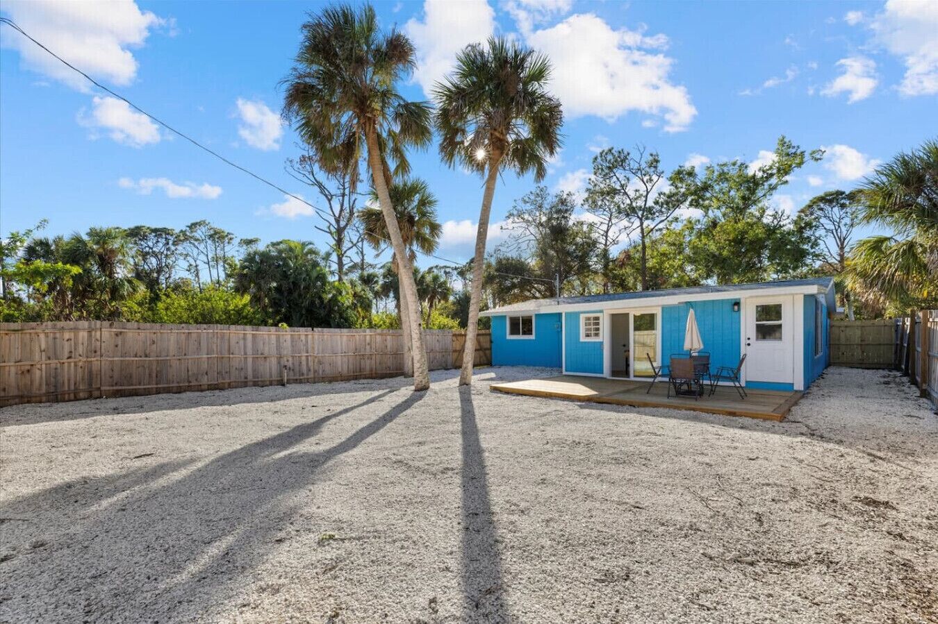 A small blue mobile home with a patio area, surrounded by a wooden fence and palm trees.