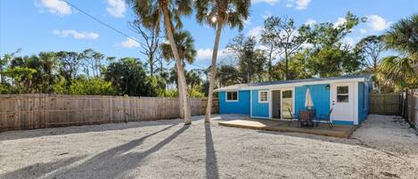 A small blue mobile home with a patio area, surrounded by a wooden fence and palm trees.