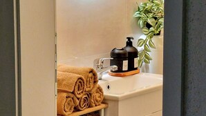 Een badkamer met een wit wastafelmeubel, een spiegel met ronde verlichting, een zwarte koffiezetapparaat en een plant.