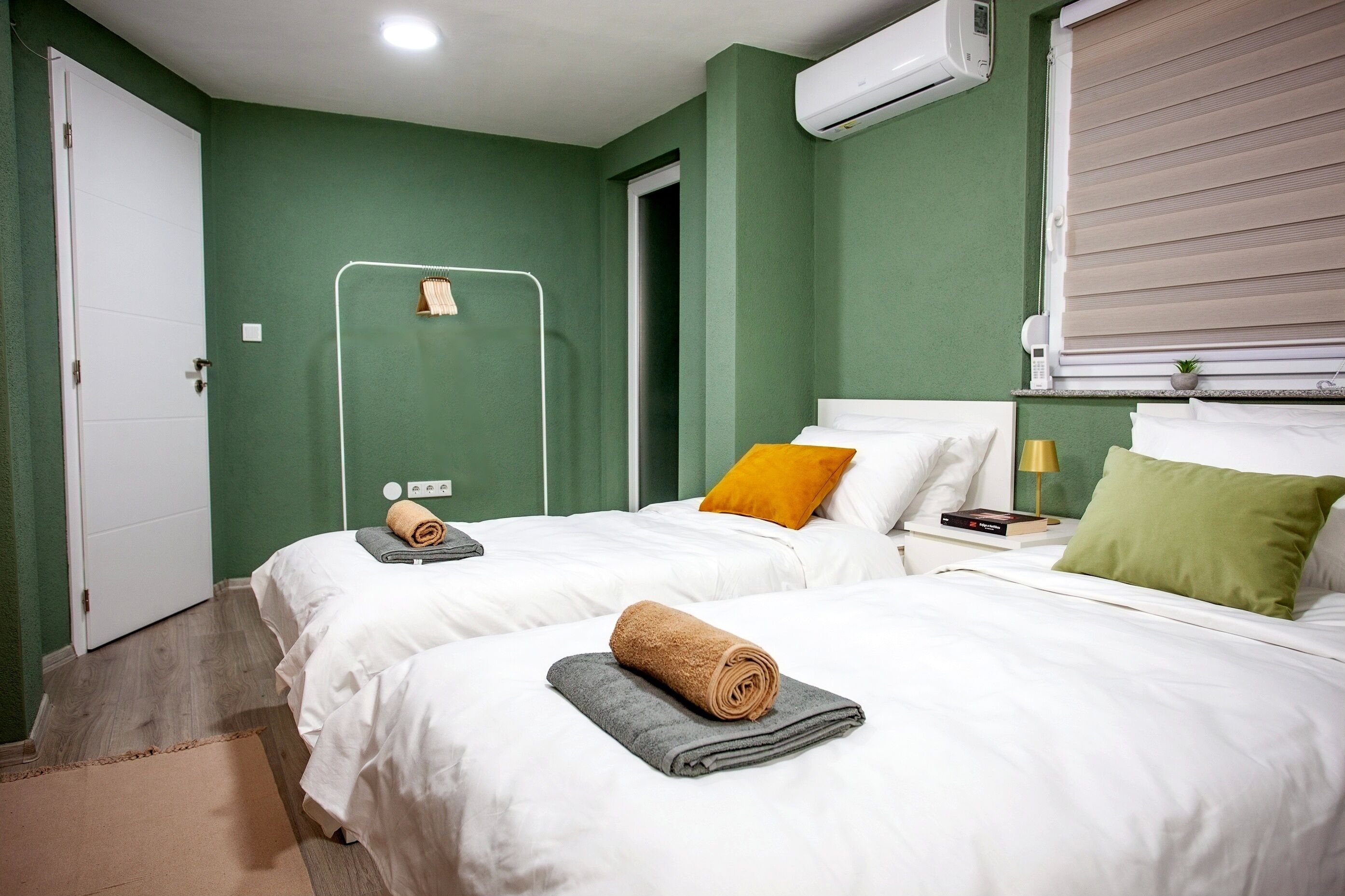 Quarto de hotel com duas camas, uma porta branca, um aparelho de ar condicionado, uma janela com persianas e uma mesa de cabeceira com abajur e um livro.