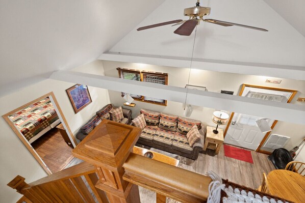 A spacious living area with a high ceiling, a ceiling fan, a sofa set, a wooden coffee table, and a staircase leading to an upper level. - Patoka Lake, Stones Throw Cabin (Birdseye)