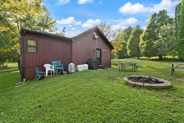 A rustic cabin with a small porch, a fire pit, and picnic tables in a grassy area surrounded by trees.