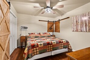 A bedroom with a wooden floor, a bed with a quilt, a ceiling fan, a wooden door, and a wall-mounted lamp. - Patoka Lake, Stones Throw Cabin (Birdseye)
