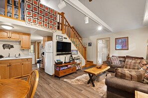 A spacious living area with a wooden staircase, a kitchen with wooden cabinets, a dining table, and a cozy seating area with a sofa and cushions.