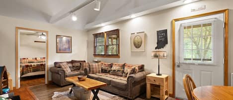 A cozy living room with a sofa, coffee table, and a dining table. There is a door with a window, a framed picture on the wall, and a rug on the floor.