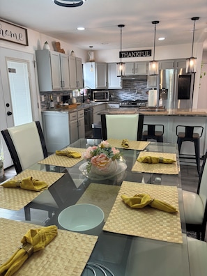 A modern kitchen with white cabinets, a stainless steel refrigerator, a microwave, and a dining area with a glass table set for six with yellow napkins.