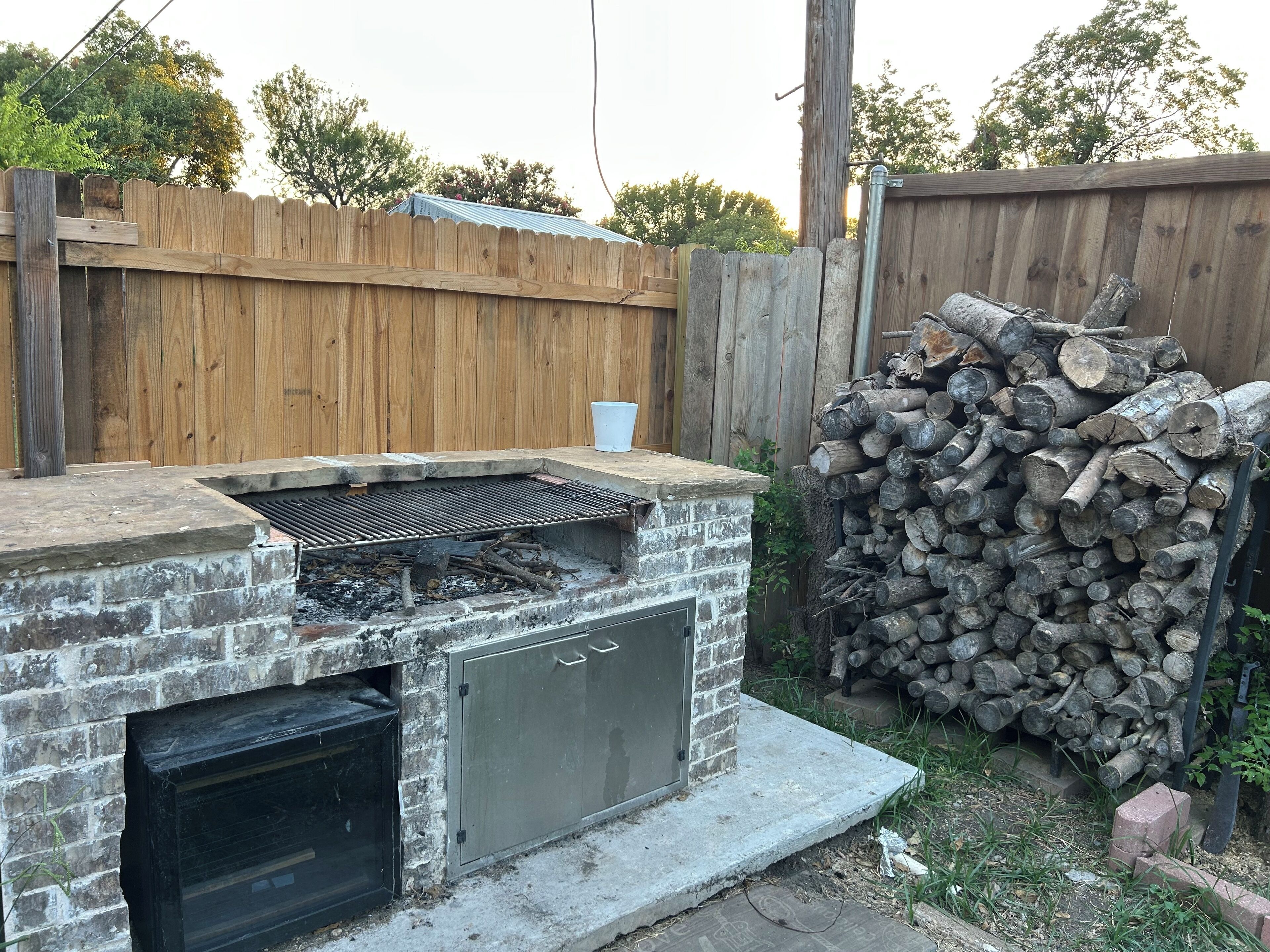 Una chimenea de ladrillo al aire libre con una rejilla metálica, rodeada por una valla de madera y una pila de leña.