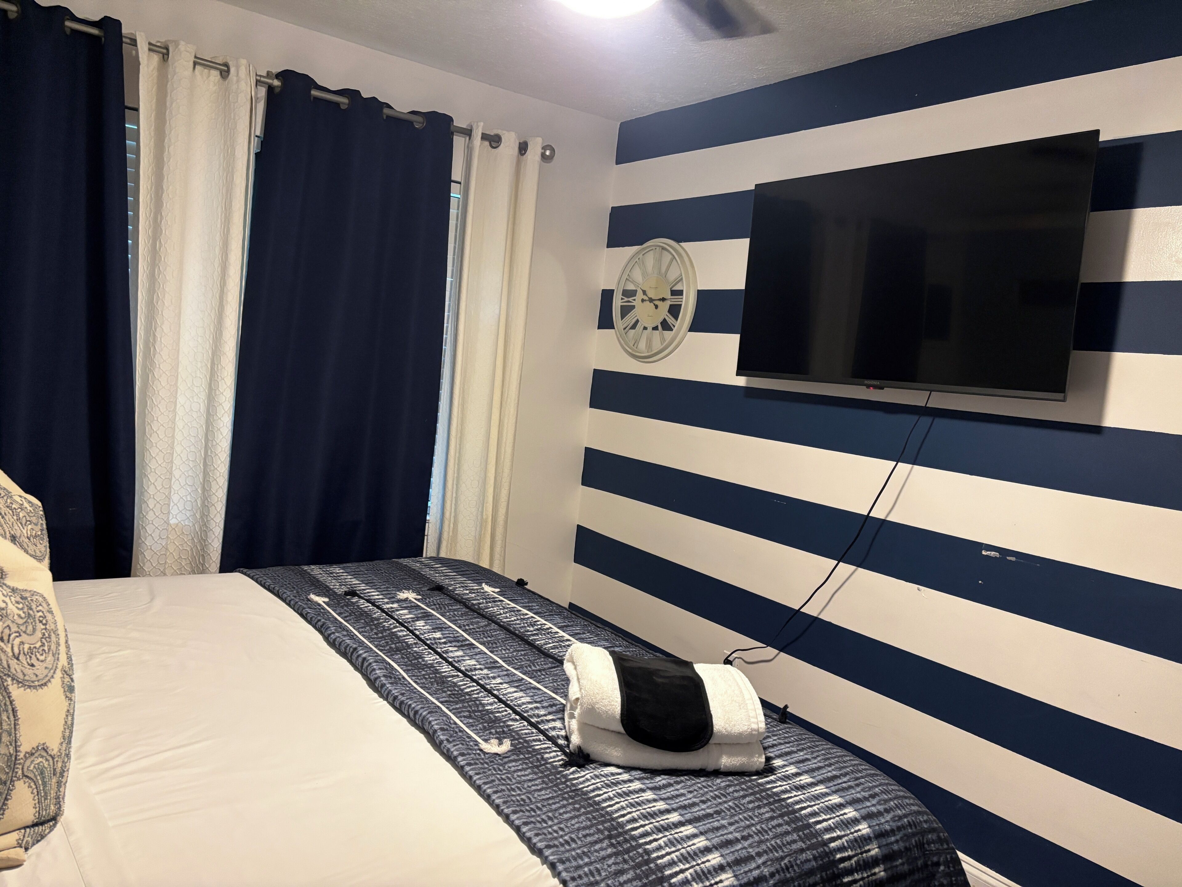 A hotel room with a bed, curtains, a wall clock, and a TV mounted on the wall.