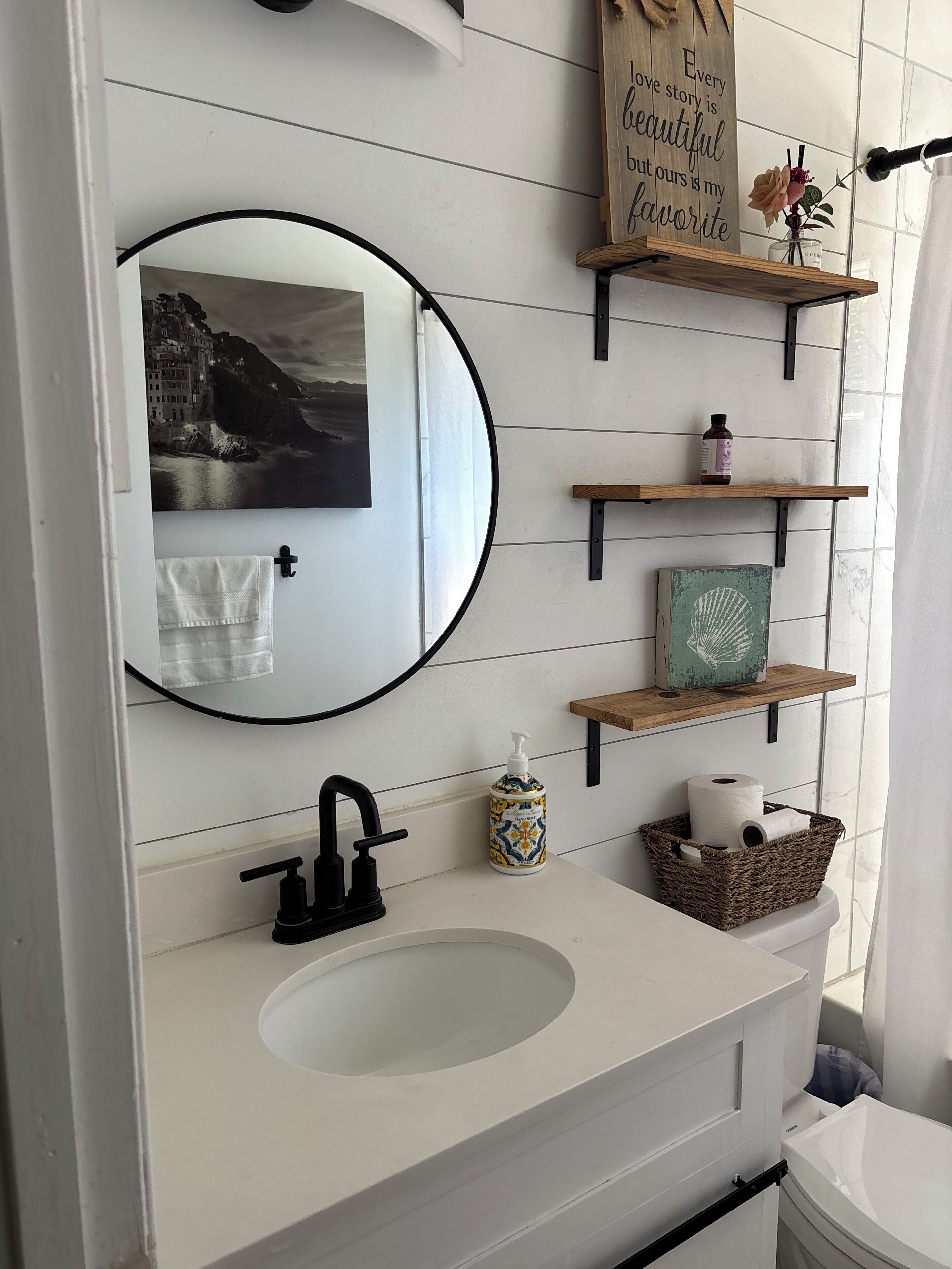 A bathroom with a white sink, black faucet, a round mirror, a framed picture, and wooden shelves with toiletries.