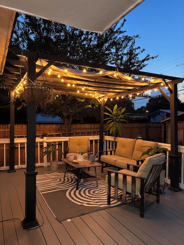 A covered outdoor patio with string lights, a cushioned seating area, and a wooden table.