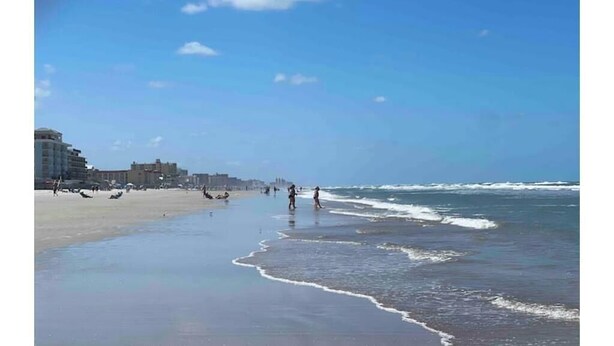 Una playa con cielo despejado, olas y edificios al fondo.