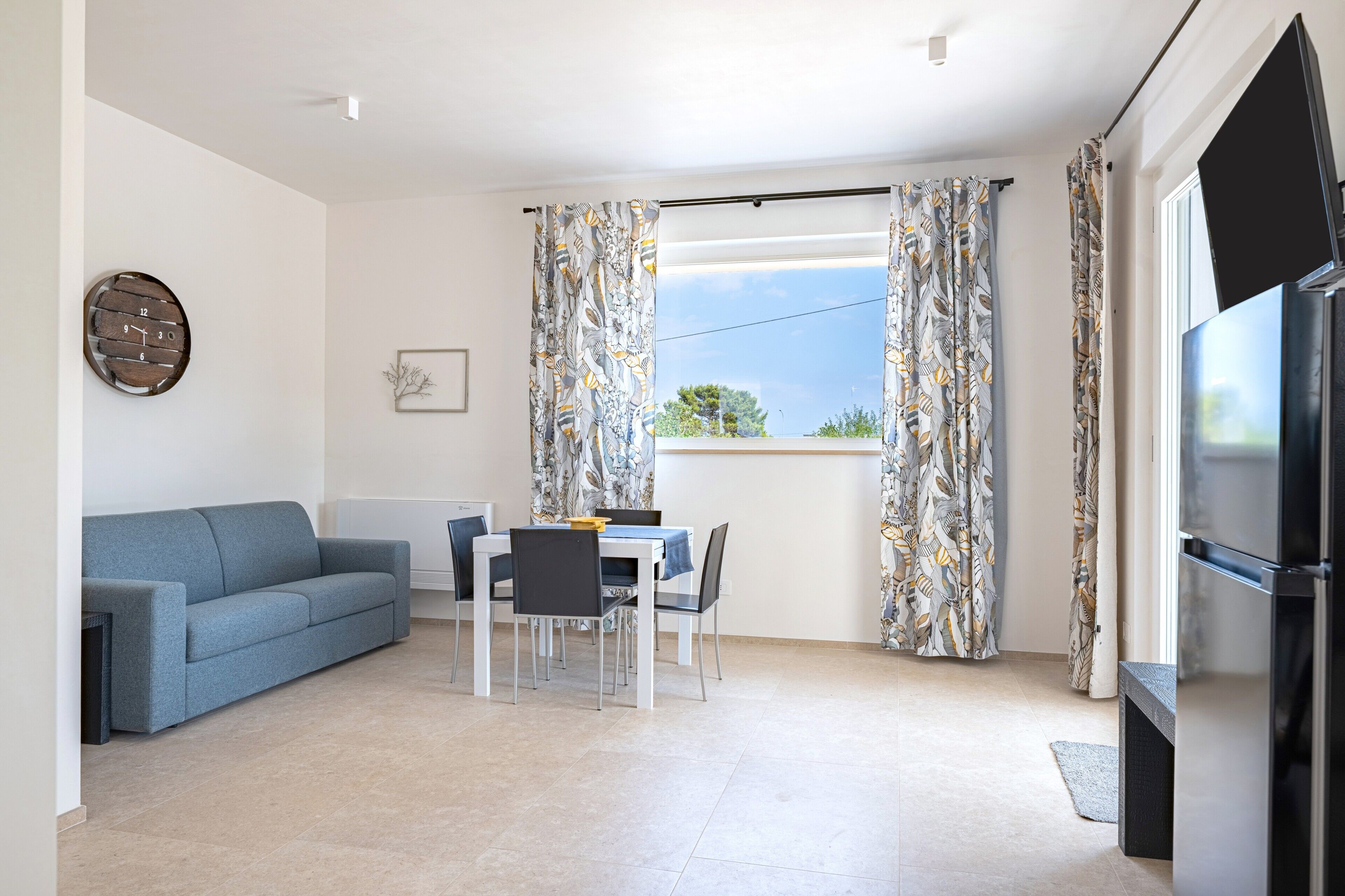 A living room with a sofa, dining table, chairs, a flat-screen TV, and a window with curtains.