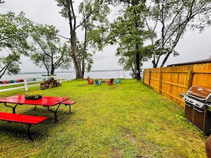A grassy backyard with a wooden fence, picnic tables, and a barbecue grill.