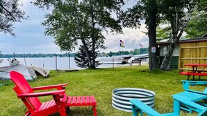 Un patio junto al lago con sillas Adirondack rojas y azules, una parrilla metálica y una terraza de madera.