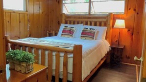 Un dormitorio de cabaña de madera con una cama, una lámpara de noche y una planta pequeña sobre un aparador.