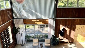 A spacious living area with a large sofa, a coffee table, and two chairs. There is a wine rack on the left and a wooden table with chairs outside the window.