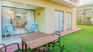 Un patio con mesa y sillas de madera, un vidrio que da a una cocina y un pequeño jardín.