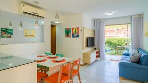 Una sala moderna con un sofá azul, una zona de comedor con sillas naranjas y un televisor sobre un mueble de madera.