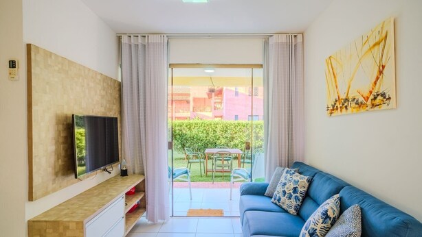 Una sala de estar moderna con un televisor de pantalla plana, un sofá azul con cojines estampados, una mesa de centro de madera y un cerramiento de vidrio corredero que da a un patio exterior con una mesa y sillas.
