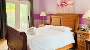 Un dormitorio con una cama de madera, dos mesitas de noche con lámparas, un ventanal con cortinas y un cuadro floral en la pared.