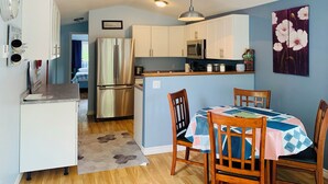 Cocina compacta con armarios blancos, un refrigerador de acero inoxidable y un microondas. Hay un comedor con una mesa y sillas, y una alfombra en el suelo.