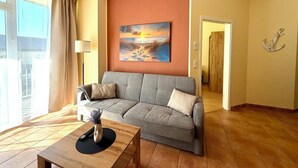 A living room with a sofa, a coffee table, a painting, and a view of the outdoors. - Südwind 05 - apartment, close to the beach, swimming pool, sauna, parking, WLAN, and balcony (Zempin)