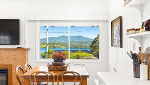 A dining area with a table and chairs, a view of a lake and mountains, a flat-screen TV, and a kitchen area with utensils and jars. - Nestle In Narooma - 10 Hillcrest Ave North Narooma (North Narooma)
