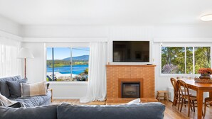 A living room with a fireplace, a sofa, a dining table with chairs, and large windows offering a view of a lake and mountains. - Nestle In Narooma - 10 Hillcrest Ave North Narooma (North Narooma)