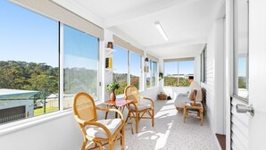 A balcony with wicker furniture, a table, and a view of greenery and buildings. - Nestle In Narooma - 10 Hillcrest Ave North Narooma (North Narooma)