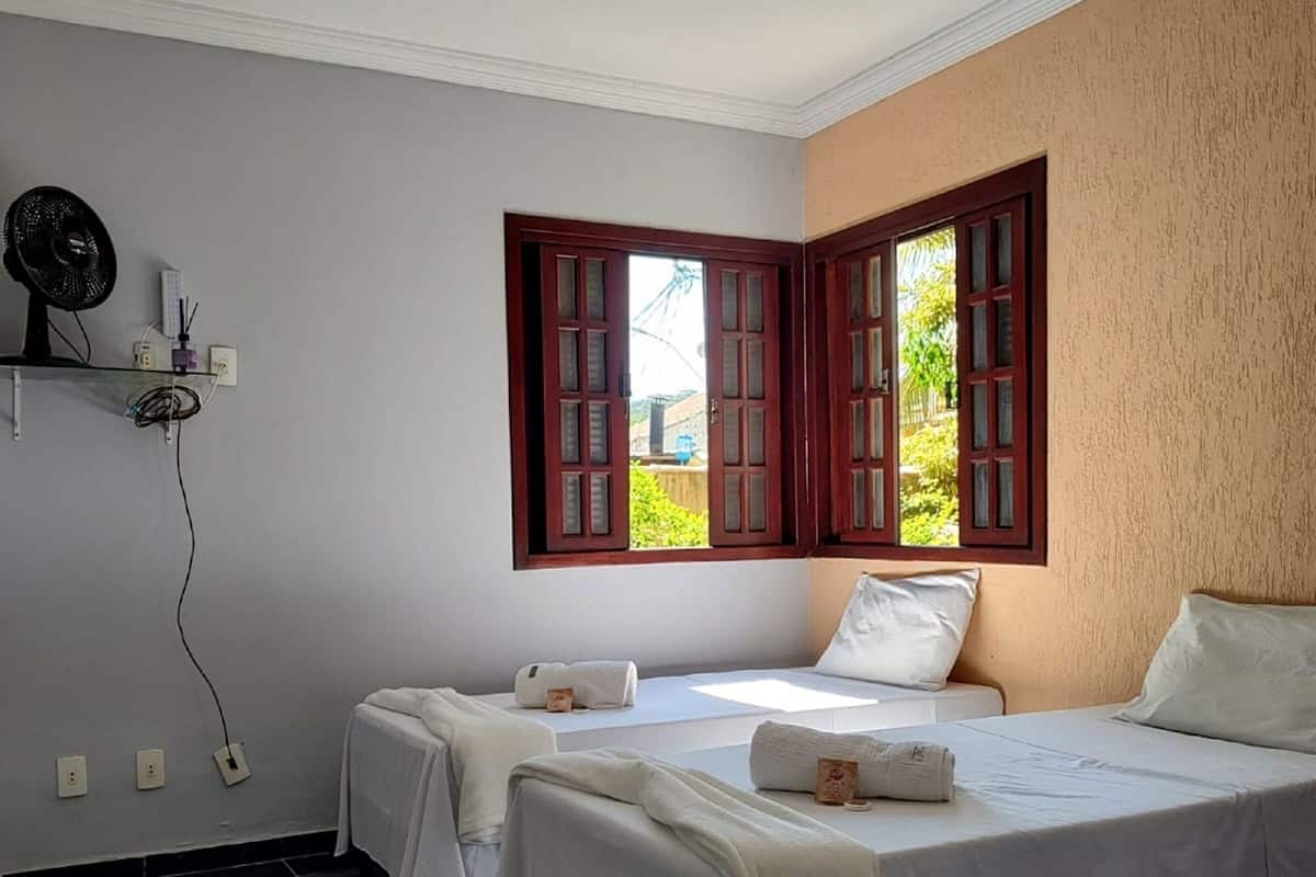 Habitación con dos camas, ventana con contraventanas rojas, ventilador de techo y un estante pequeño con un ventilador y un control remoto.