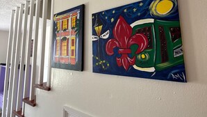 A hallway with white walls and a white ceiling. There are two paintings on the wall, one depicting a building and the other a green bus with a red flower.