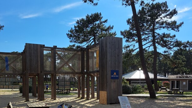 A wooden play structure with a sign reading "AIRE DE JEUX" in a sandy area with picnic tables and benches. - Mobilhome 6 pers (Vendays-Montalivet)
