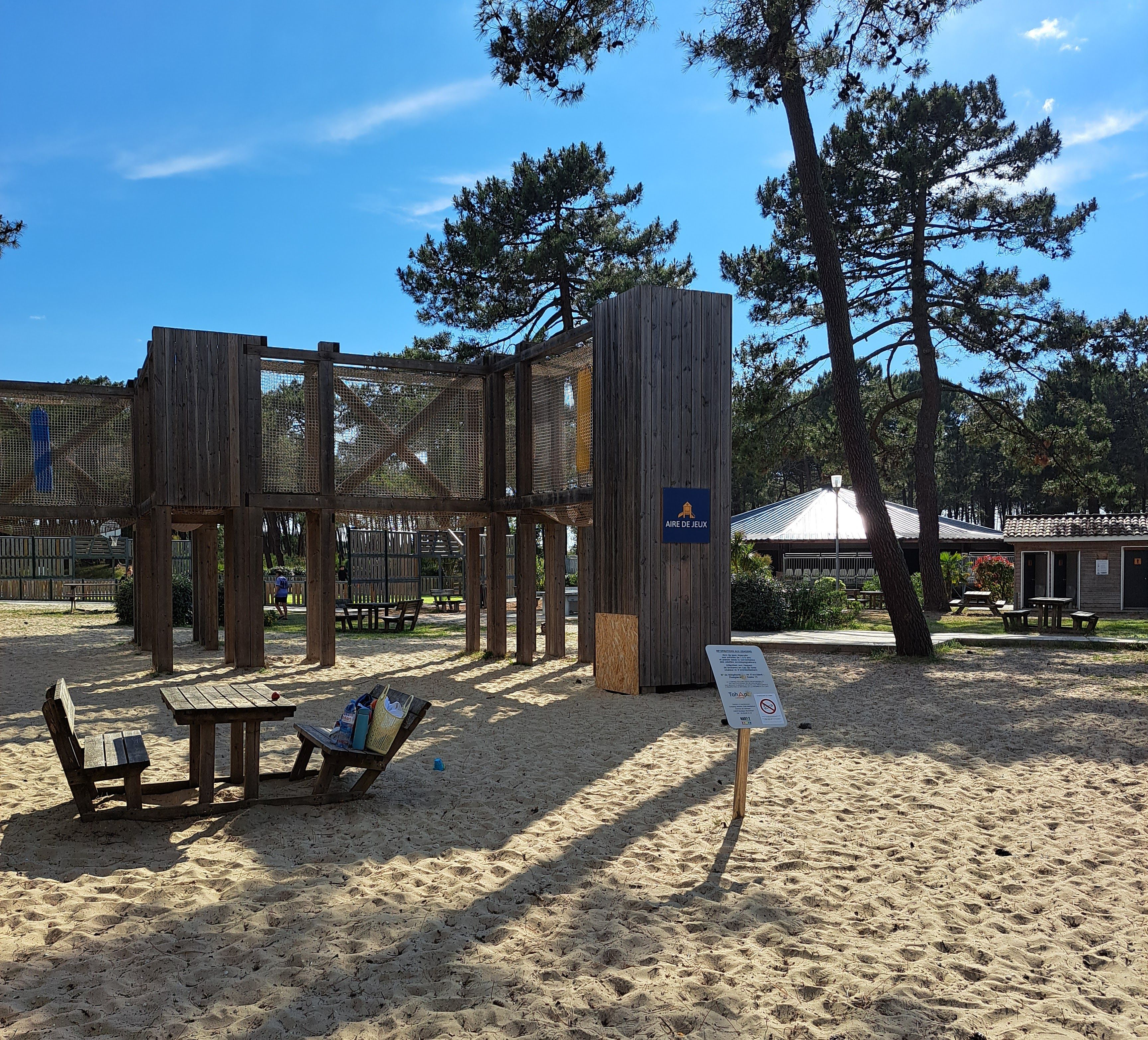 A wooden play structure with a sign reading "AIRE DE JEUX" in a sandy area with picnic tables and benches.