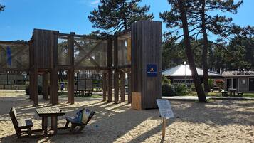 A wooden play structure with a sign reading "AIRE DE JEUX" in a sandy area with picnic tables and benches.