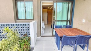 Un balcon avec une table en bois et des chaises bleues, une vue sur une pièce où se trouve un lit, et un petit espace jardin avec des plantes.