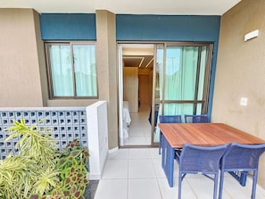 Un balcon avec une table en bois et des chaises bleues, une vue sur une pièce où se trouve un lit, et un petit espace jardin avec des plantes.