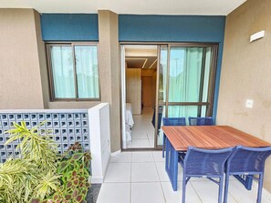 A balcony with a wooden table and blue chairs, a view into a room with a bed, and a small garden area with plants. - Makia - Porto de Galinhas  (Ipojuca)