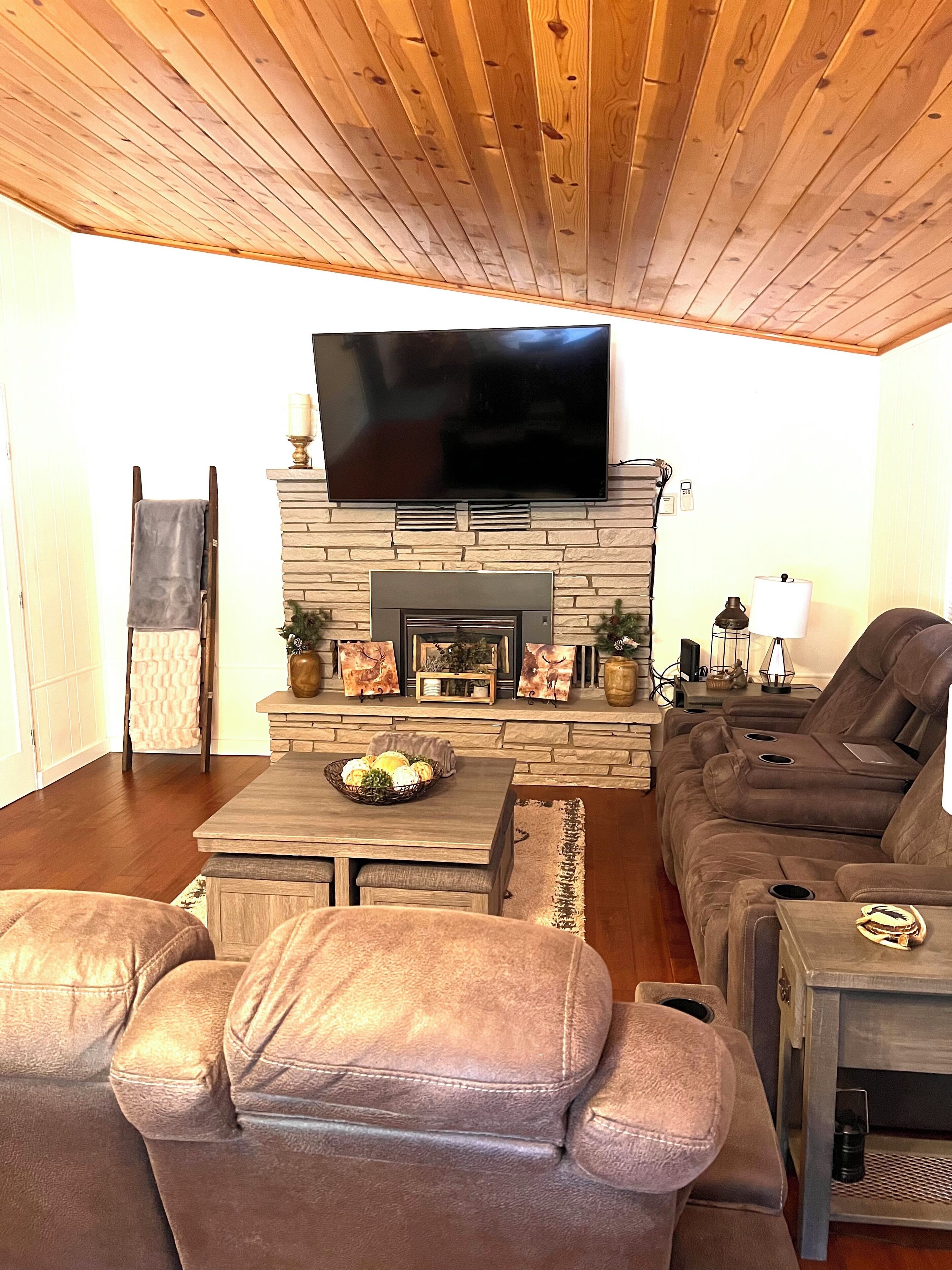 A living room with a stone fireplace, a flat-screen TV, leather sofas, and a wooden ceiling.