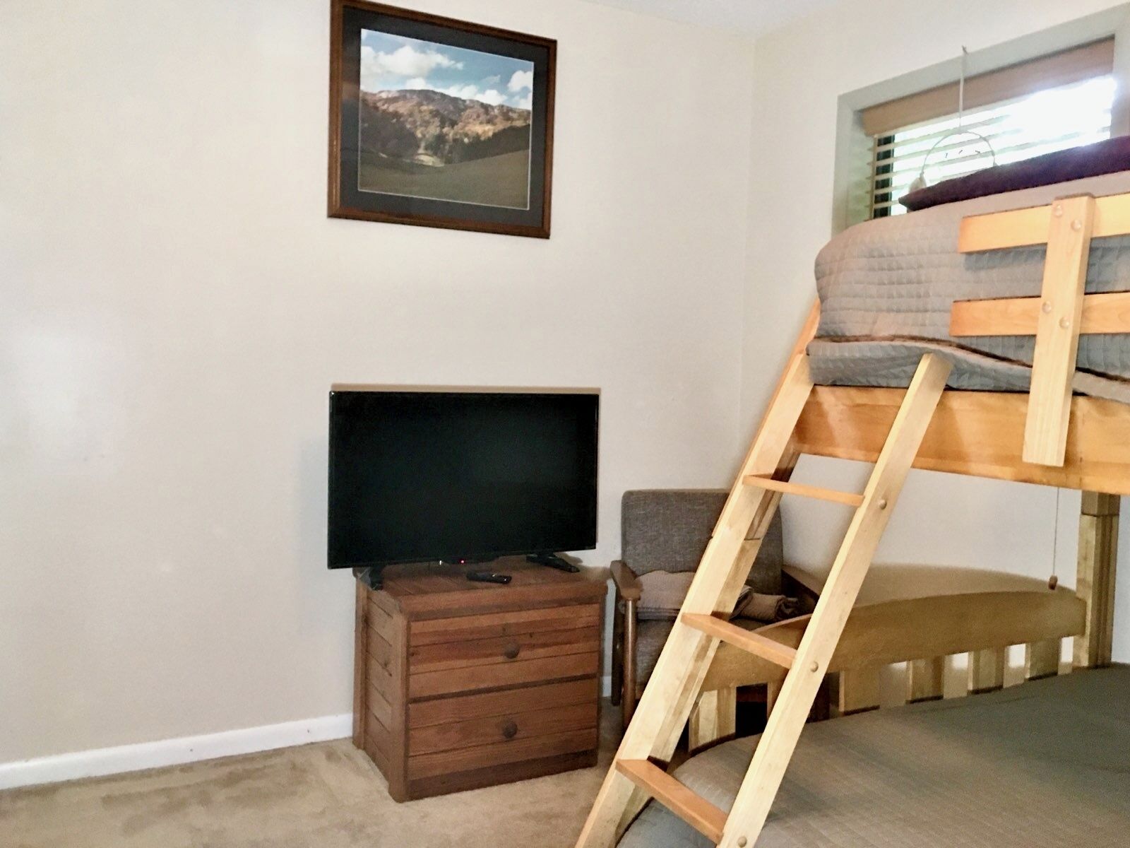 Un dormitorio con una cama litera, un televisor sobre un aparador de madera, un cuadro enmarcado en la pared y una ventana con persianas.