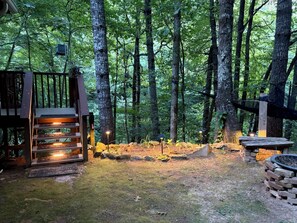 A forested area with a wooden deck, stone steps, and a fire pit.