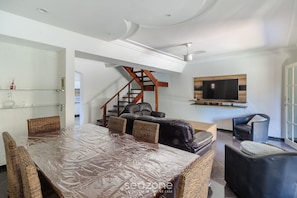 A spacious living area with a large dining table, comfortable seating, a staircase, and a mounted TV.