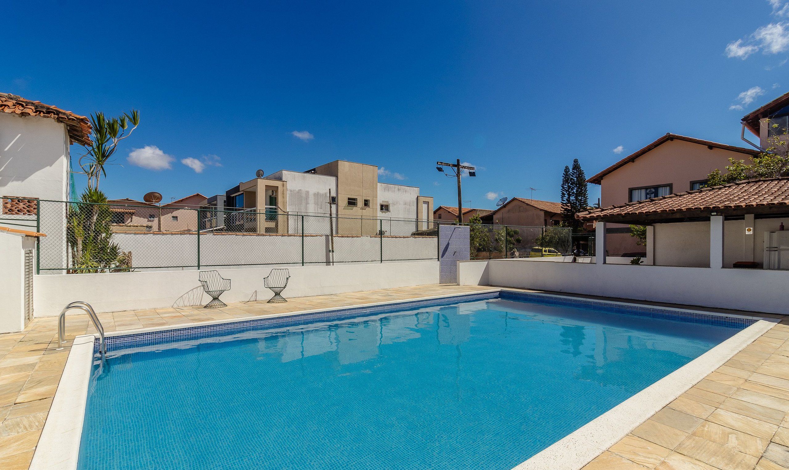 A swimming pool with a tiled deck, surrounded by buildings with a fence, and a clear blue sky.