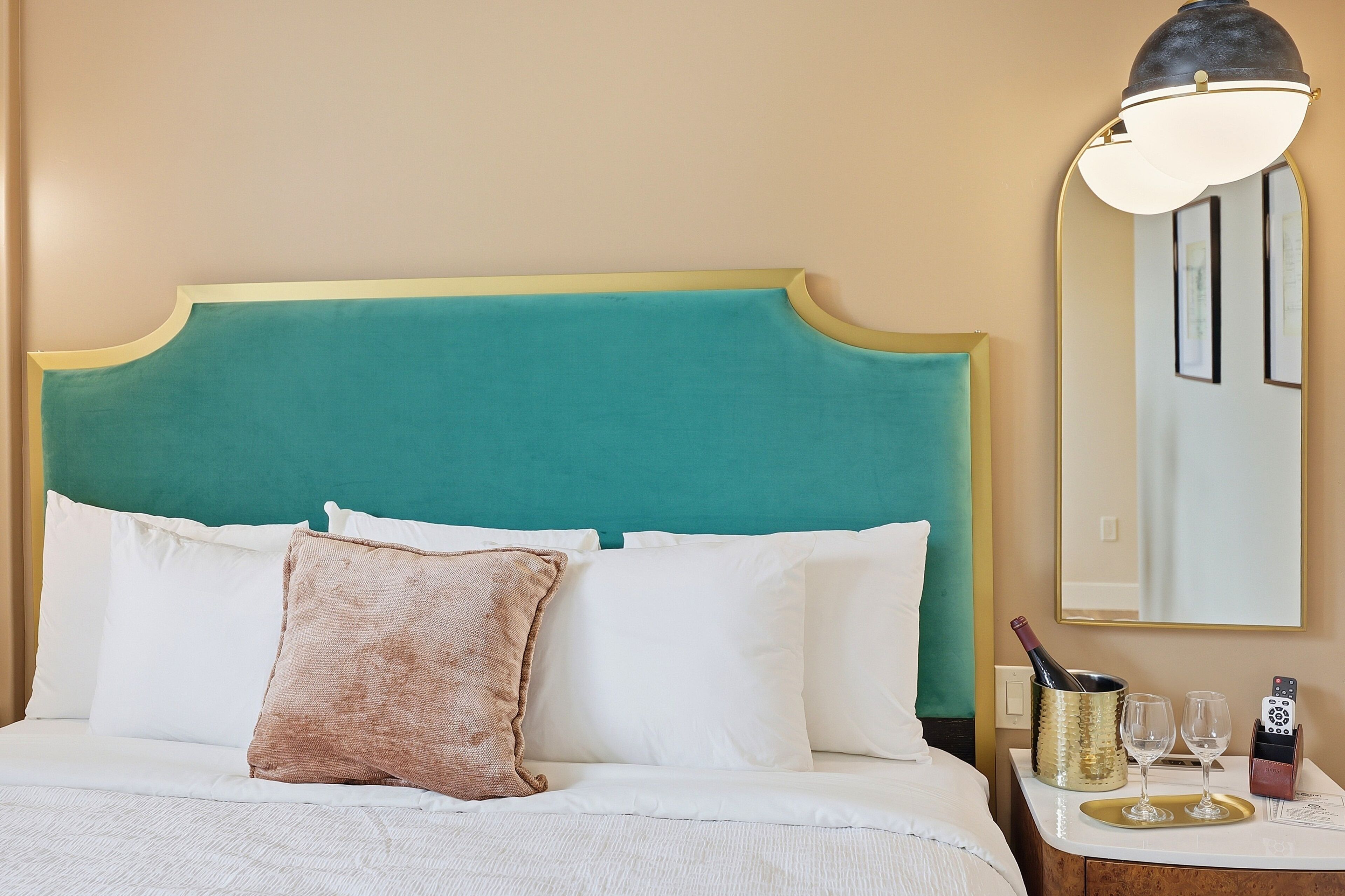 A hotel room with a teal headboard, white bedding, a brown pillow, a bedside table with a bottle, glasses, and a remote control.