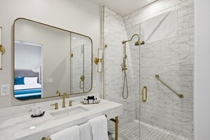 Une salle de bain moderne avec un grand miroir, une douche équipée d’un pommeau de douche à main et un lavabo blanc avec des accessoires dorés.