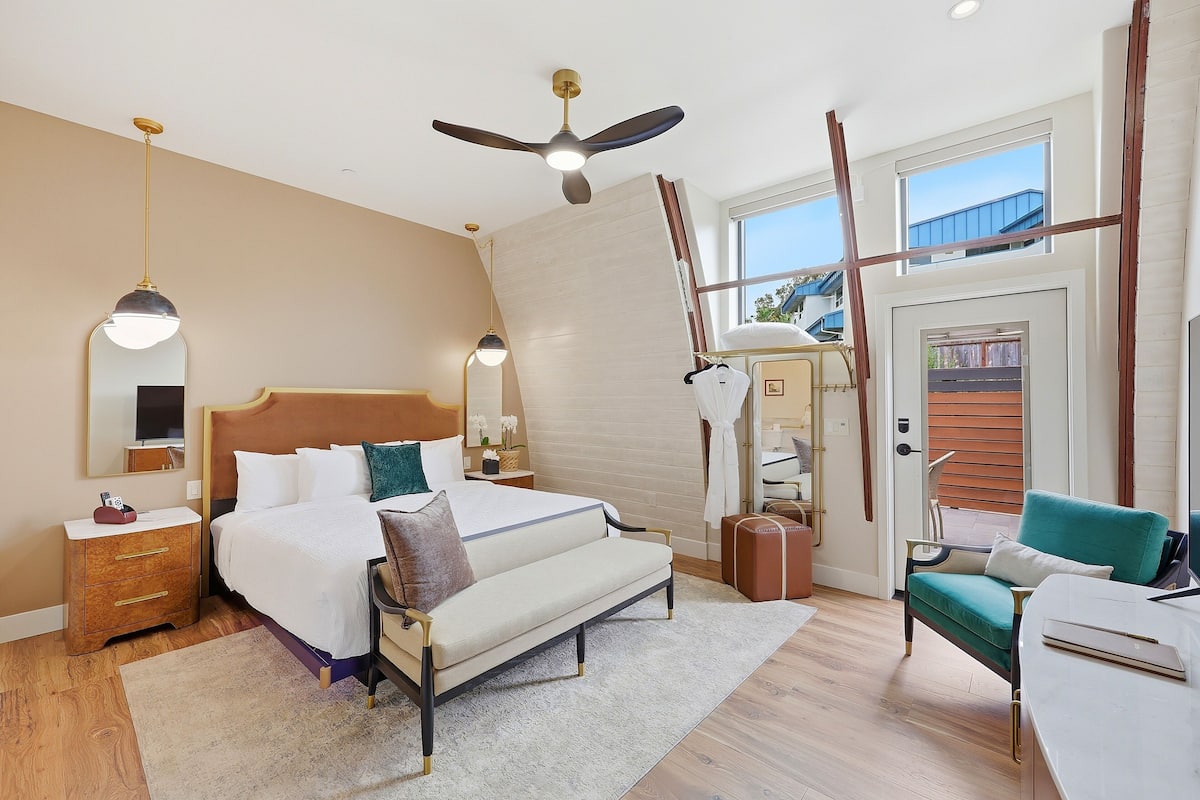 A hotel room with a bed, bedside tables, a desk, a chair, a ceiling fan, and a large window.
