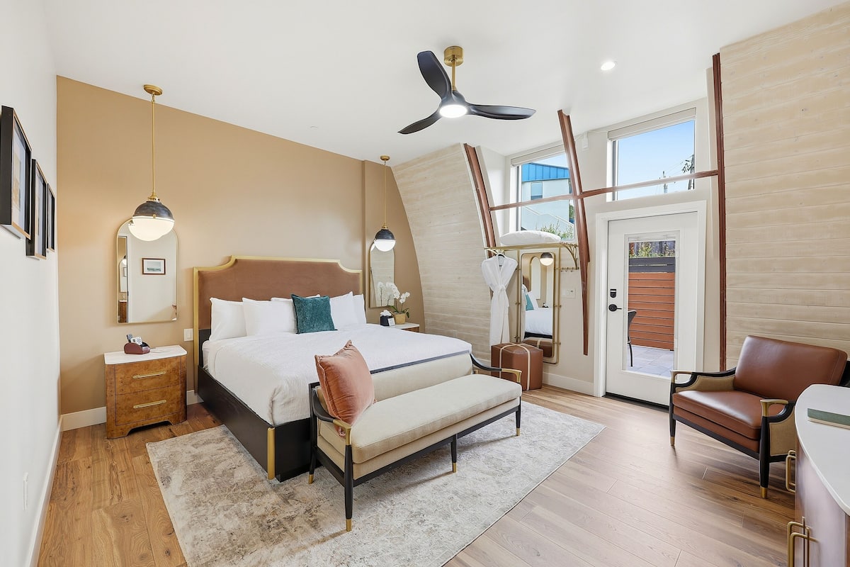 A bedroom with a bed, bedside tables, a bench, a chair, a ceiling fan, and a large window.