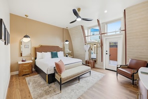 A bedroom with a bed, bedside tables, a bench, a chair, a ceiling fan, and a large window. - The Q Inn (Morro Bay)