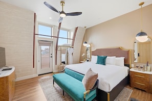 Une chambre d’hôtel moderne avec un grand lit, un ventilateur de plafond, un banc, une table de chevet avec une lampe et une grande fenêtre.