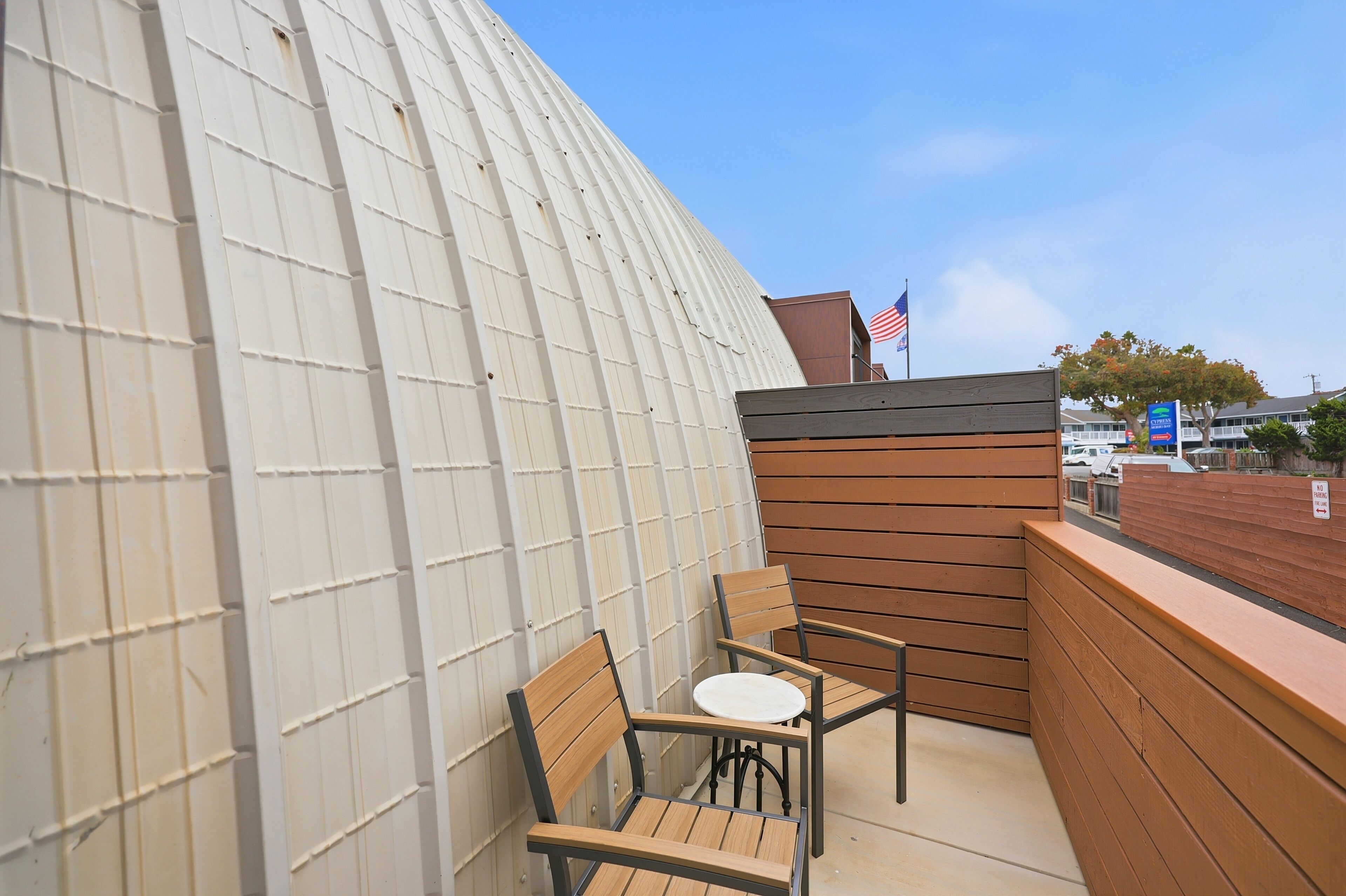 A balcony with two wooden chairs and a small table, a corrugated metal wall, and a flag.